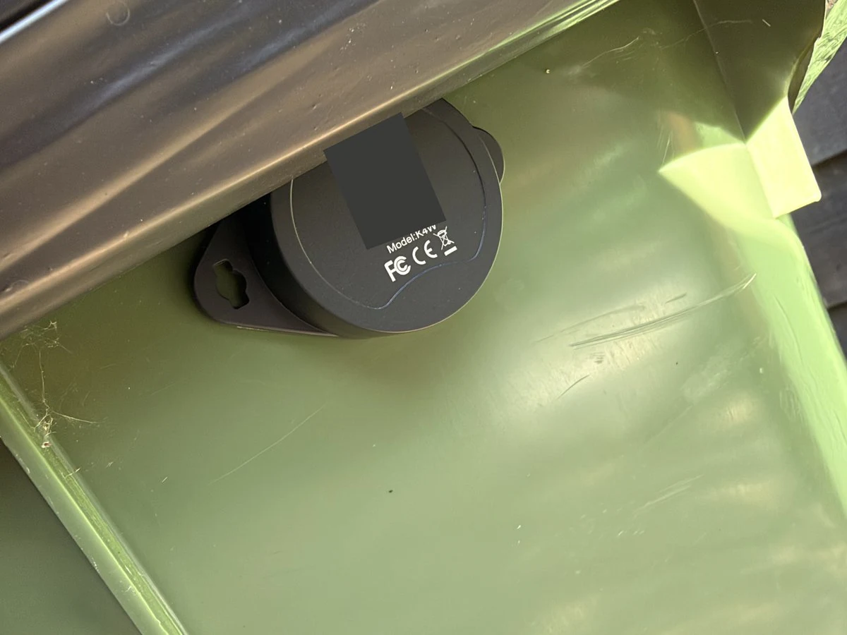 A KKM K4W tag fixed beneath the rim of a green wheelie bin, with its identifying label obscured.